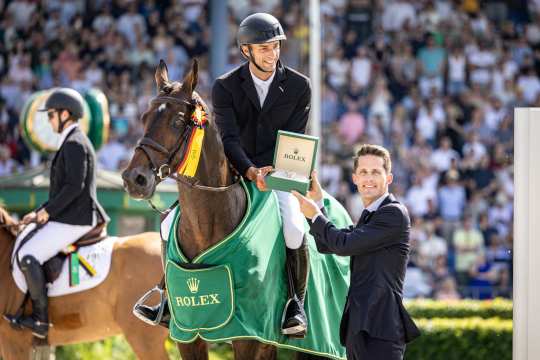 Sieger des Rolex Grand Prix auf seinem Pferd erhält bei der Siegerehrung eine Rolex-Uhr, umgeben von weiteren Reitern und dicht gedrängtem Publikum im Stadion.