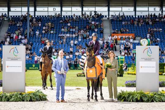 Siegerehrung beim CHIO Aachen in einem Springreitstadion: Eine Reiterin auf einem braunen Pferd mit orangefarbener Siegerdecke der Handwerkskammer Aachen steht zwischen zwei grauen Tafeln mit dem Logo „Weltfest des Pferdesports CHIO Aachen“. Links sitzt ein weiterer Reiter auf seinem Pferd, vorne stehen zwei Offizielle mit Pokal und Blumen. Im Hintergrund sind die Tribünen mit vielen Zuschauern zu sehen.