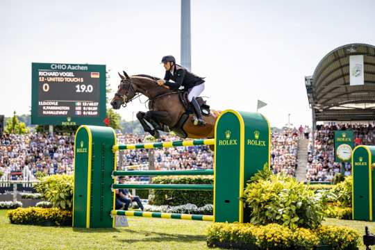 A show jumper wearing a black jacket and helmet jumps over a green-and-yellow Rolex obstacle on a bay horse in a packed stadium; on the left is a scoreboard displaying “CHIO Aachen.”