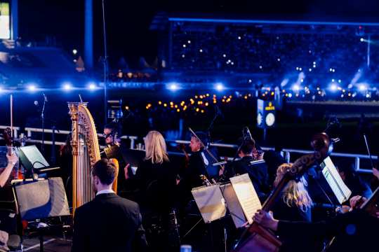Ein Orchester spielt im Vordergrund mit Harfe und Streichern, während im Hintergrund das Reitstadion in blauem Licht leuchtet; in der Arena sind viele kleine Feuerlichter zu sehen.