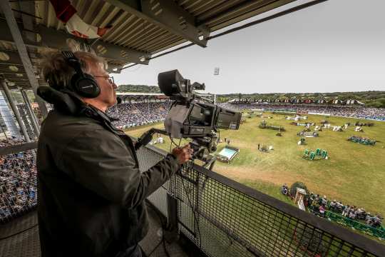 Kameramann mit großen Kopfhörern filmt mit einer professionellen TV-Kamera von einer überdachten Tribüne aus ein gut besuchtes Reitstadion; unten auf dem weitläufigen Rasenplatz sind Hindernisse und Parcours-Elemente aufgebaut, rundherum sitzen zahlreiche Zuschauer auf den Rängen.