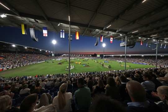 Panoramic view of a fully packed equestrian stadium at dusk: beneath the roof with many national flags, thousands of spectators watch a brightly lit grass arena set up with multiple show jumping fences.