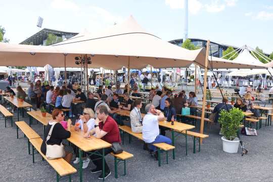 Besucher sitzen an langen Holztischen unter einem großen Sonnensegel; im Hintergrund stehen Messe- und Gastronomiestände auf dem Veranstaltungsgelände.