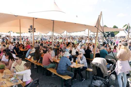 Unter großen Sonnensegeln sitzen viele Besucher an langen Bierbänken und Tischen; es ist ein belebter Gastrobereich bei sonnigem Wetter, im Vordergrund steht ein Kinderwagen.