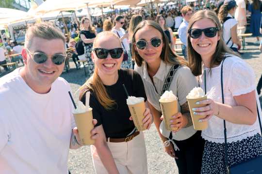 Vier junge Erwachsene lächeln in die Kamera und halten auf einem sonnigen Eventgelände große Becher mit Sahnegetränken; im Hintergrund sind viele Besucher und Sonnenschirme zu sehen.