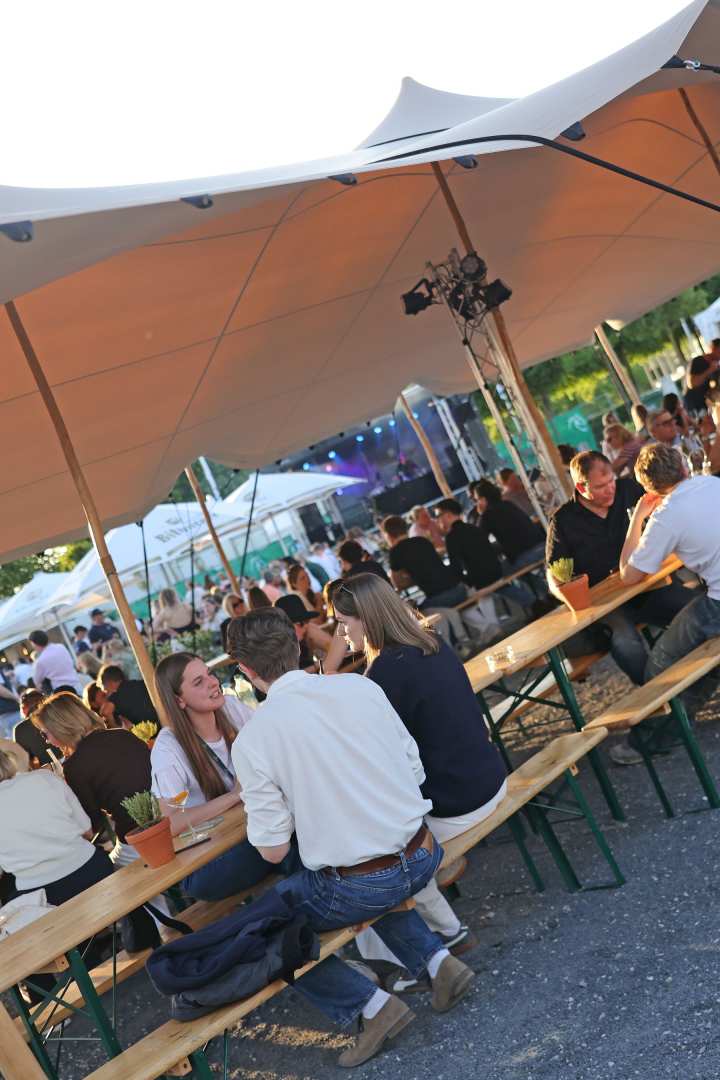 Besucher sitzen am Abend unter einem großen Sonnensegel an langen Holzbänken und unterhalten sich; im Hintergrund leuchtet eine Bühne mit farbigem Licht.