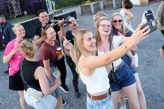 A group of young adults is taking a selfie at an event venue; some are holding beer and wine glasses, and in the background stands a musician with a violin.