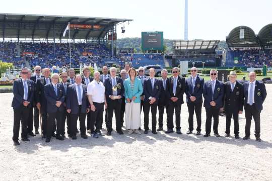 Gruppenfoto im Reitstadion: Viele Männer in dunklen Sakkos mit Vereinsabzeichen stehen neben einer Frau im türkisfarbenen Blazer; im Hintergrund sind Tribünen und eine Anzeigetafel zu sehen.
