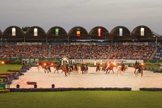 Abendliche Stadionaufnahme: Viele Reiter auf Pferden bewegen sich durch die Arena, am Rand leuchten Scheinwerfer; die Tribüne ist voll besetzt und unter dem Dach hängen Nationalflaggen, an der Bande steht „Rolex Grand Slam of Show Jumping“.