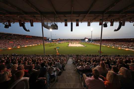 Weitwinkelaufnahme eines abendlichen Reitstadions, dicht gefüllte Tribünen im Vordergrund, im hell ausgeleuchteten Rasenoval in der Mitte eine sandfarbene Dressurfläche mit mehreren Pferden und Reitern, umgeben von Flutlichtmasten und Länderflaggen unter dem Stadiondach.