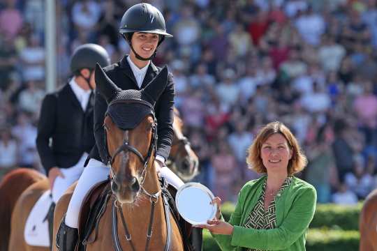 Eine Springreiterin in schwarzem Sakko und Helm sitzt auf einem braunen Pferd im Stadion und lächelt; daneben hält eine Frau im grünen Blazer einen runden, silbernen Teller in die Kamera, im Hintergrund ist Publikum unscharf zu sehen.