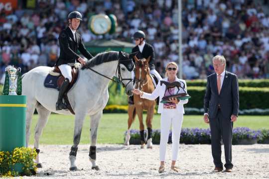 Ein Springreiter in schwarzem Sakko sitzt auf einem Schimmel im Stadion; daneben hält eine Frau in Weiß eine Pferdefigur-Trophäe, rechts steht ein Mann im Anzug, im Hintergrund sind Publikum und ein großer Rolex-Uhrenaufbau zu sehen.