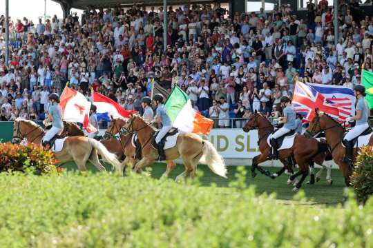 Mehrere Reiter reiten im Stadion an einer voll besetzten Tribüne vorbei und tragen große Nationalflaggen; im Hintergrund ist ein „Rolex Grand Slam“-Banner zu sehen.