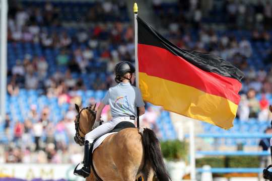Eine Reiterin in hellblauem Shirt und Helm reitet im Stadion und trägt eine große deutsche Flagge; im Hintergrund sind Zuschauer auf der Tribüne unscharf zu sehen.