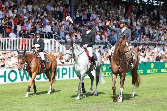 Drei Reiter reiten im Stadion an einer voll besetzten Tribüne vorbei und winken mit weißen Tüchern; im Hintergrund sind grüne Rolex-Banden zu sehen.