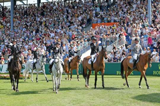 Mehrere Reiter reiten im Stadion vor einer voll besetzten Tribüne und winken mit weißen Tüchern; im Hintergrund sind grüne Rolex-Banden und ein orangefarbenes STAWAG-Banner zu sehen.