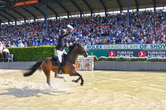 Ein Reiter in schwarzem Sakko reitet mit einem braunen Pferd durch ein Wasserhindernis im Stadion und hält ein weißes Tuch hoch; im Hintergrund jubelt eine voll besetzte Tribüne mit „Rolex Grand Slam“- und „Turkish Airlines“-Banden.