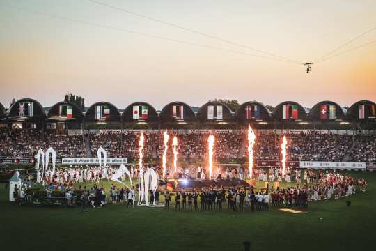 Weitwinkelaufnahme der Eröffnungsfeier des CHIO Aachen 2025 im voll besetzten Reitstadion: Vor den Tribünen mit zahlreichen Nationalflaggen stehen Menschen auf dem Rasen, während mehrere Feuerfontänen aufsteigen.