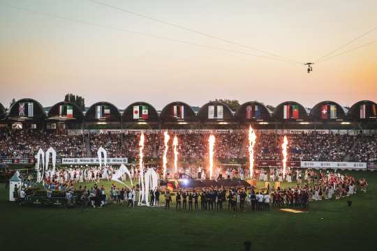 Weitwinkelaufnahme der Eröffnungsfeier des CHIO Aachen 2025 im voll besetzten Reitstadion: Vor den Tribünen mit zahlreichen Nationalflaggen stehen Menschen auf dem Rasen, während mehrere Feuerfontänen aufsteigen.